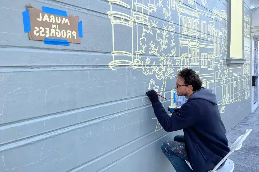 An artist paints a mural on the side of a building in the Mission District, 在建筑上贴了一个牌子，上面写着“壁画正在进行中”.加州贝博体彩app.